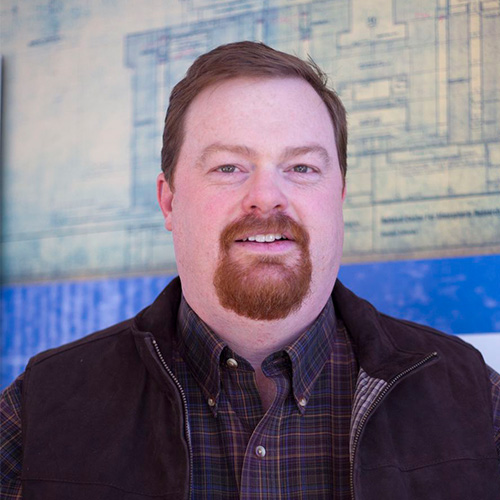 Headshot of National Renewable Energy researcher Aaron Andersen. He smiles and wears a black plaid button up with black jacket. He stands in front of a piece of cream and blue art.