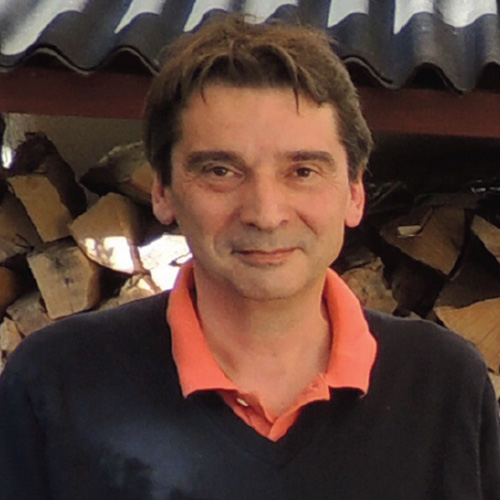 Headshot of BNL researcher Alexei Klimentov. He smiles and wears a dark colored pull over with a coral colored undershirt. He stands against a pile of wood. 