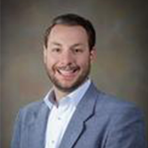 Headshot of Sandia researcher Andrew Younge. He smiles and wears a light colored button up with gray blazer. He stands against a brown backdrop.