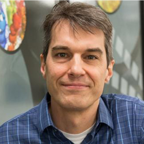 Headshot of Argonne researcher Tom Uram. He smiles and wears a blue flannel button up. He stands in front of a piece of art.