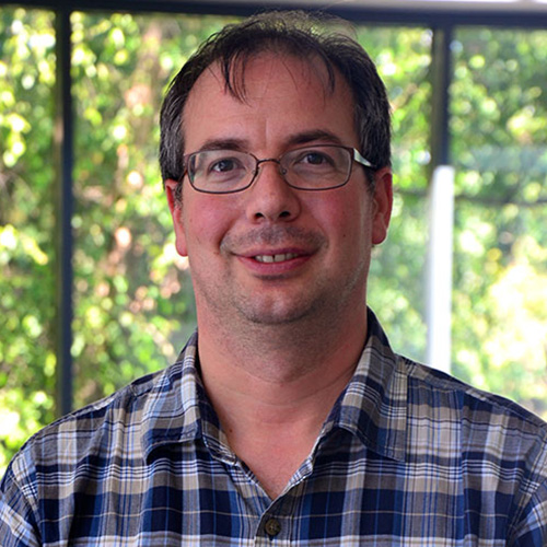 Headshot of JLAB researcher Balint Joo. He smiles and wears a blue striped button up. He stands against a nature background.