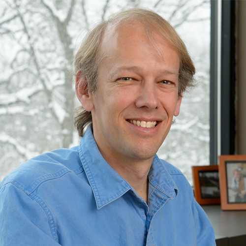 Headshot of Argonne researcher Pete Beckman. He smiles, wears a light blue button up, and sits in his office. Photos and a snow covered tree can be seen in the background. 