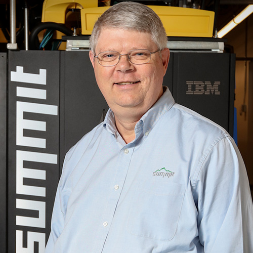 Photo of Oak Ridge researcher Arthur "Buddy" Bland. He smiles and wears a 'Summit' blue button up. He stands next to the Summit Super Computer.