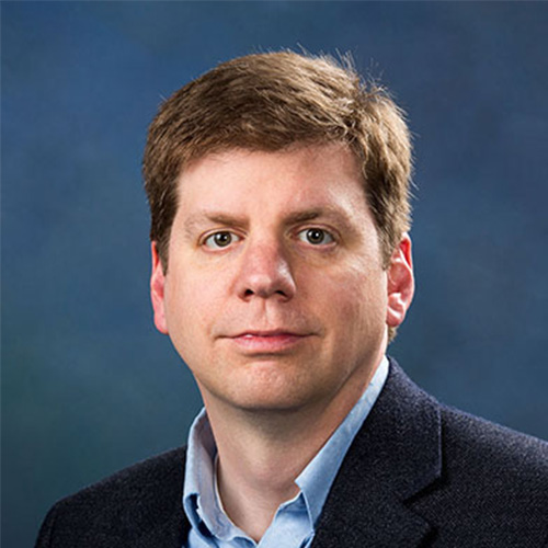 Headshot of Los Alamos researcher Brad Settlemyer. He smiles and wears a blue button up with black blazer. He stands against a blue backdrop.