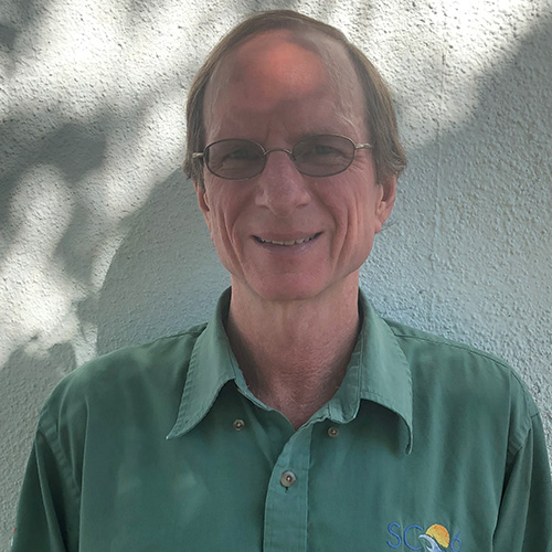 Headshot of Sandia researcher Jim Brandt. He smiles and wears a light green button up with a super computing embroidery detail. He stands against a white background.