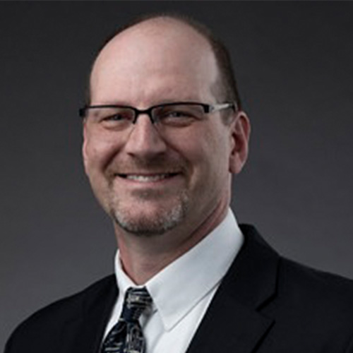 Headshot of Los Alamos researcher Brian Albright. He smiles and wears a white button up with black blazer. He stands against a grey backdrop.