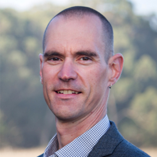 Photo of Lawrence Livermore researcher Brian Spears. He smiles and wears a blue and white button up with blazer. He stands against a nature backdrop.