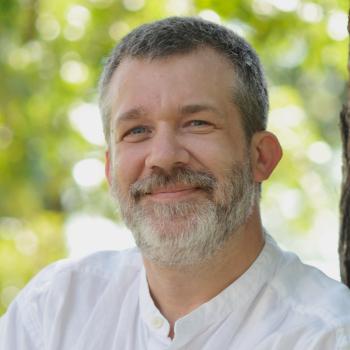 Headshot of Argonne researcher Charlie Catlett. He smiles and wears a white button up with a mock collar. He stands in front of a blurred nature scene.