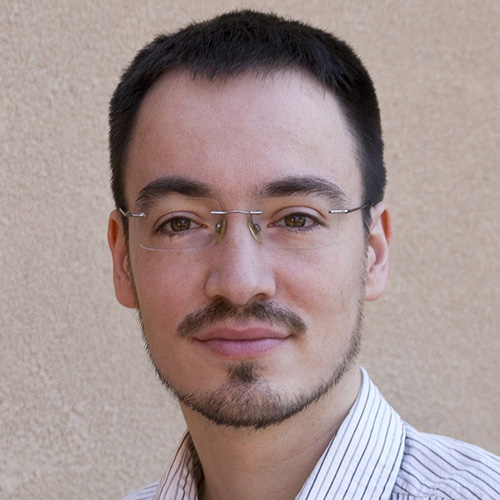 Headshot of Sandia researcher Christian Trott. He smiles and wears a white button up with stripes. He stands in front of a cream colored wall.