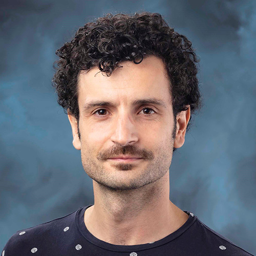 Headshot of Oak Ridge researcher Damien Lebrun. He smiles and wears a patterned top. He stands in front of a blue backdrop.