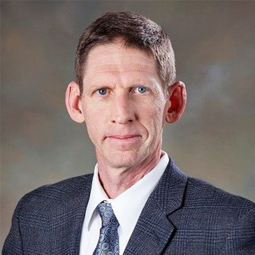 Photo of Oak Ridge researcher David Womble. He smiles and wears a white button up with blazer and a patterned blue tie. He stands against a gray backdrop.