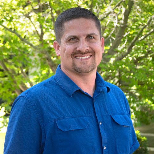 Headshot of Lawrence Livermore researcher Chris DePrater. He smiles and wears a blue button up. He stands outside in front of a tree.