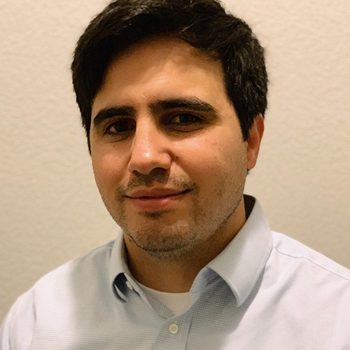 Headshot of Los Alamos researcher Dominic Manno. He smiles and wears a white button up with light blue lines. He stands in front of a white background.