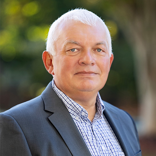 Headshot of Jefferson Lab researcher Graham Heyes. He smiles and wears a light colored button up with a navy blazer. He stands in front of a blurred nature scene.