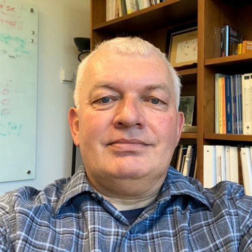 Headshot of JLAB researcher Graham Heyes. He smiles and wears a blue striped button up. He stands in his office. Books and an in-progress whiteboard can be seen in the background.