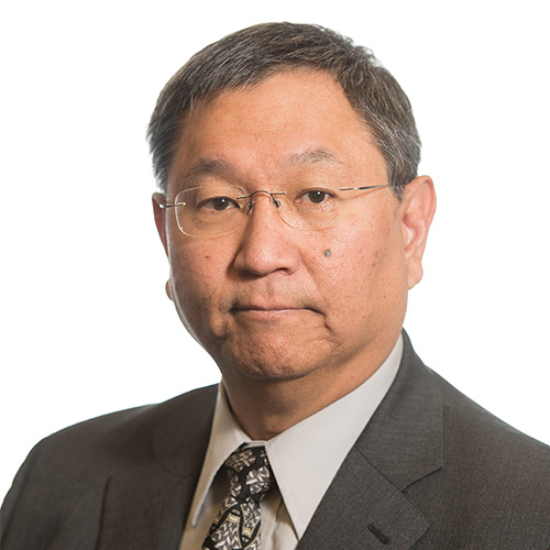 Headshot of Pacific Northwest researcher James A. Ang. He smiles and wears a gray suite with a cream and black printed tie. He stands in front of a white background.