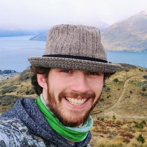 Photo of Brookhaven researcher Johannes Doerfert. He smiles and in front of a nature scene with a large body of water, mountains, and a lush valley. 