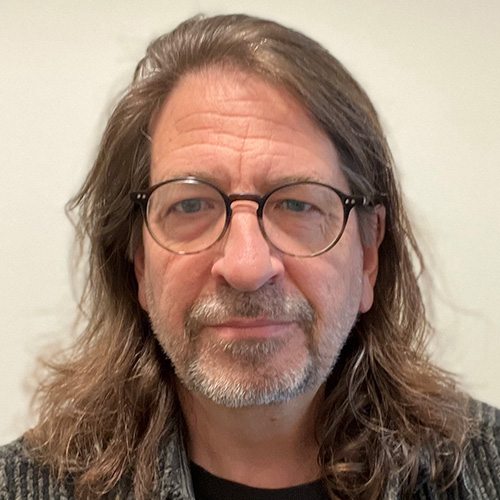 Headshot of Argonne researcher Rick Stevens. He smiles and wears a gray sweater. He has glasses. He stands in front of a cream backdrop.