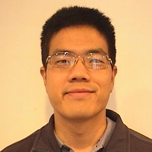 Headshot of Sandia researcher Stephen Lin He smiles and wears a brown sweater. He has glasses. He stands in front of a cream backdrop.