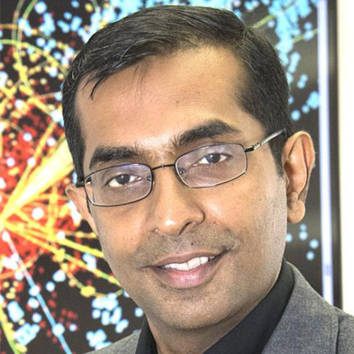 Headshot of Brookhaven researcher Shantenu Jha. He smiles and wears a gray suite with a black button up. He stands in front of a vivid colored background.