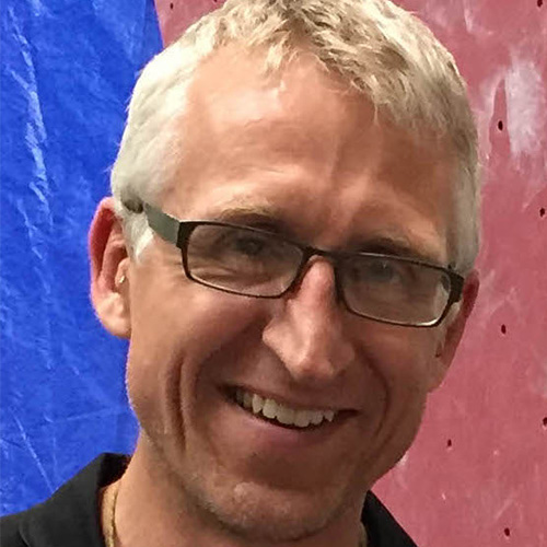 Headshot of National Renewable Energy researcher Mike Sprague. He smiles and wears glasses. He stands against a blue and red background.