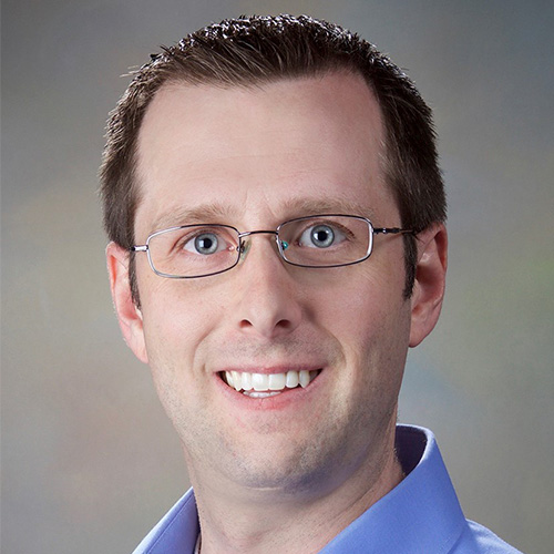 Headshot of Sandia researcher Stan Moore. He smiles and wears a blue button up. He has glasses. He stands in front of a gray backdrop.