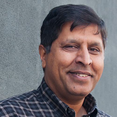 Headshot of Lawrence Berkeley researcher Sudip Dosanjh. He smiles and wears a navy flannel button up. He stands in front of a light gray background.