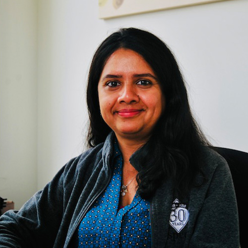 Headshot of Brookhaven researcher Sunita Chandrasekaran. She smiles and wears a blue patterned blouse with pull over. She sits in an office. A piece of wall art can be seen behind her. 