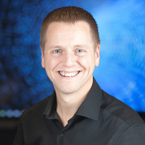 Headshot of Oak Ridge researcher Trevor Wood. He smiles and wears a black button up. He stands in front of a vivid blue backdrop.