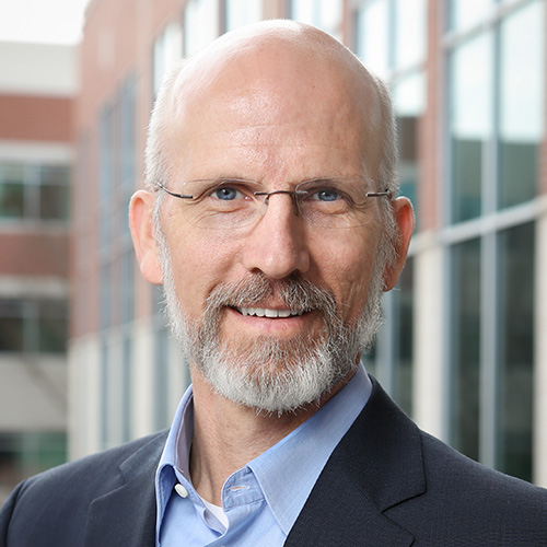 Headshot of Oak Ridge researcher Jack Wells. He smiles, wears a light colored button up with blazer. He stands against a nature scene. 