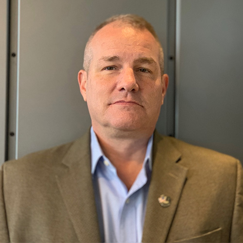 Headshot of Sandia researcher Jim Laros. He smiles and wears a light colored button up with brown blazer. He stands against a gray background.