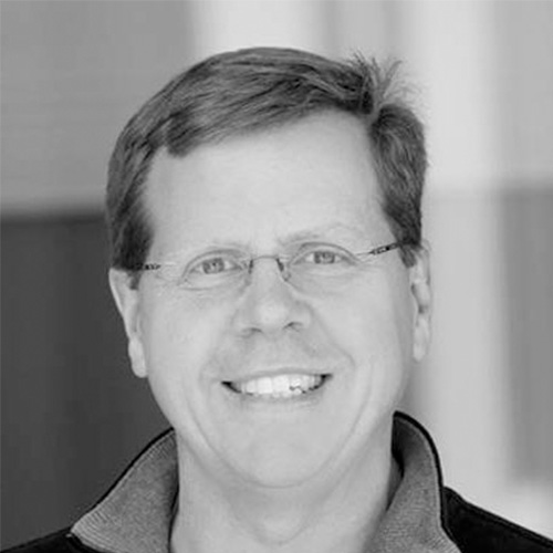 Headshot of Sandia National researcher Michael Heroux. His headshot is in black and white. He smiles and wears glasses. He stands in front of a blurred background.