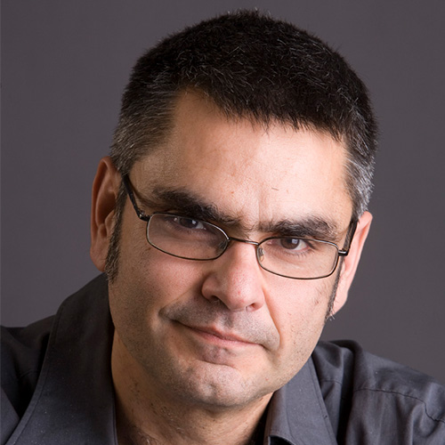 Headshot of Fermi National researcher Panagiotis Spentzouris. He smiles, has glasses, and stands against a dark grey background. 